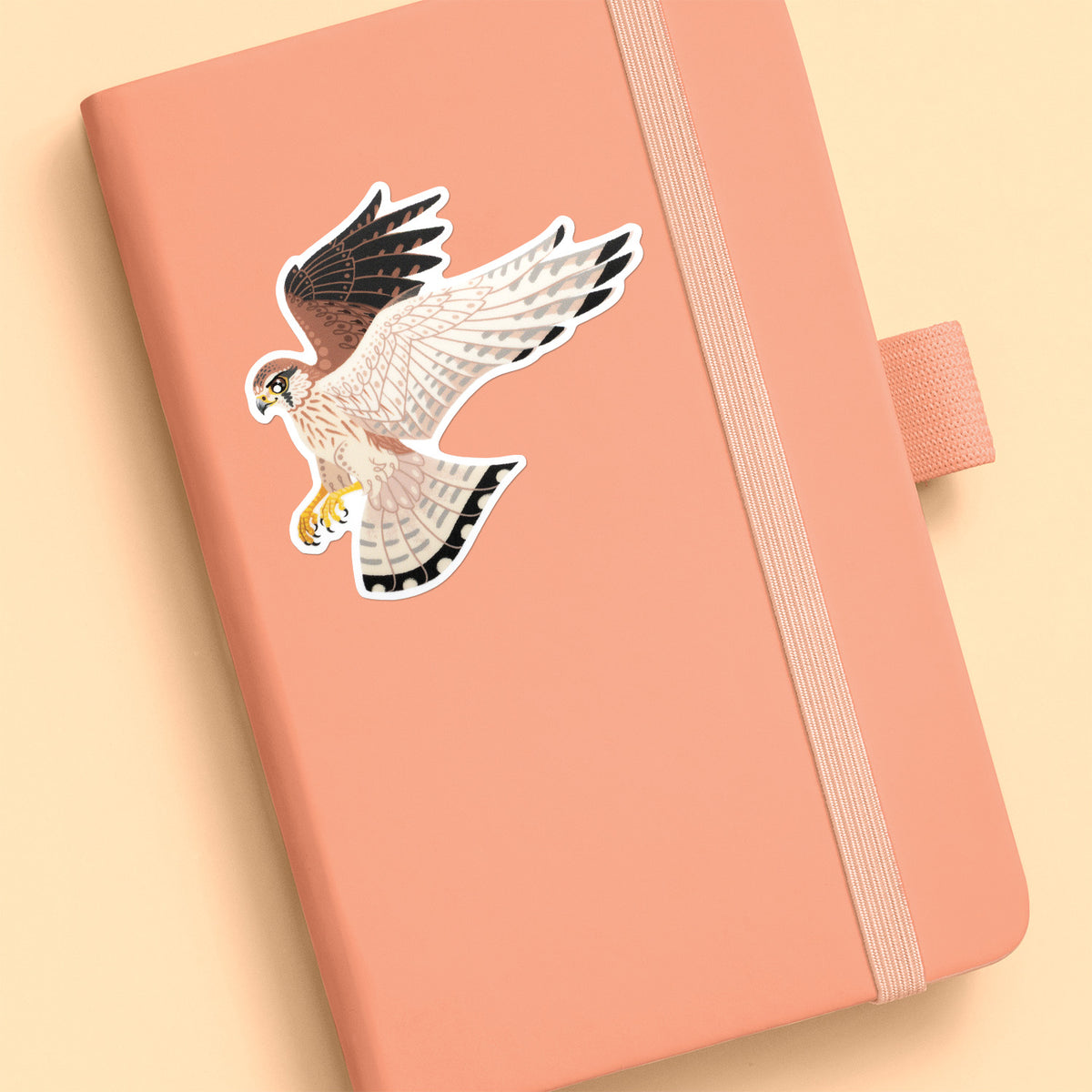Colourful sticker of a hovering female nankeen kestrel stuck to a pale orange journal on an soft yellow background.