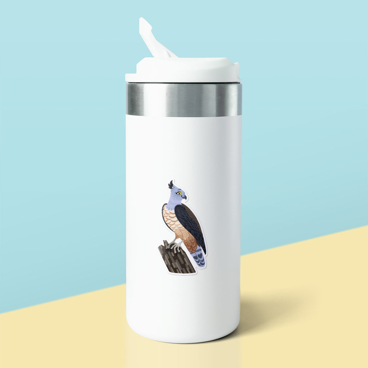 Colourful sticker of a pacific baza perched on a tree branch stuck to a white metal drink bottle on a soft yellow table on a pale teal background.