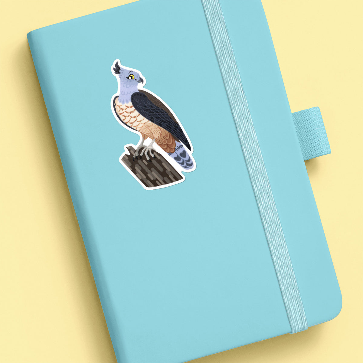 Colourful sticker of a pacific baza perched on a tree branch stuck to a teal journal on a soft yellow background.