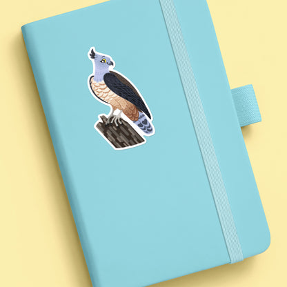 Colourful sticker of a pacific baza perched on a tree branch stuck to a teal journal on a soft yellow background.