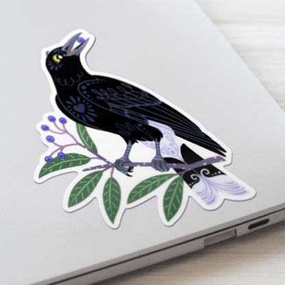 Closeup of a colourful diecut sticker of a pied currawong eating a blueberry ash berry stuck to a lid of a Macbook laptop on a textured white wood background.