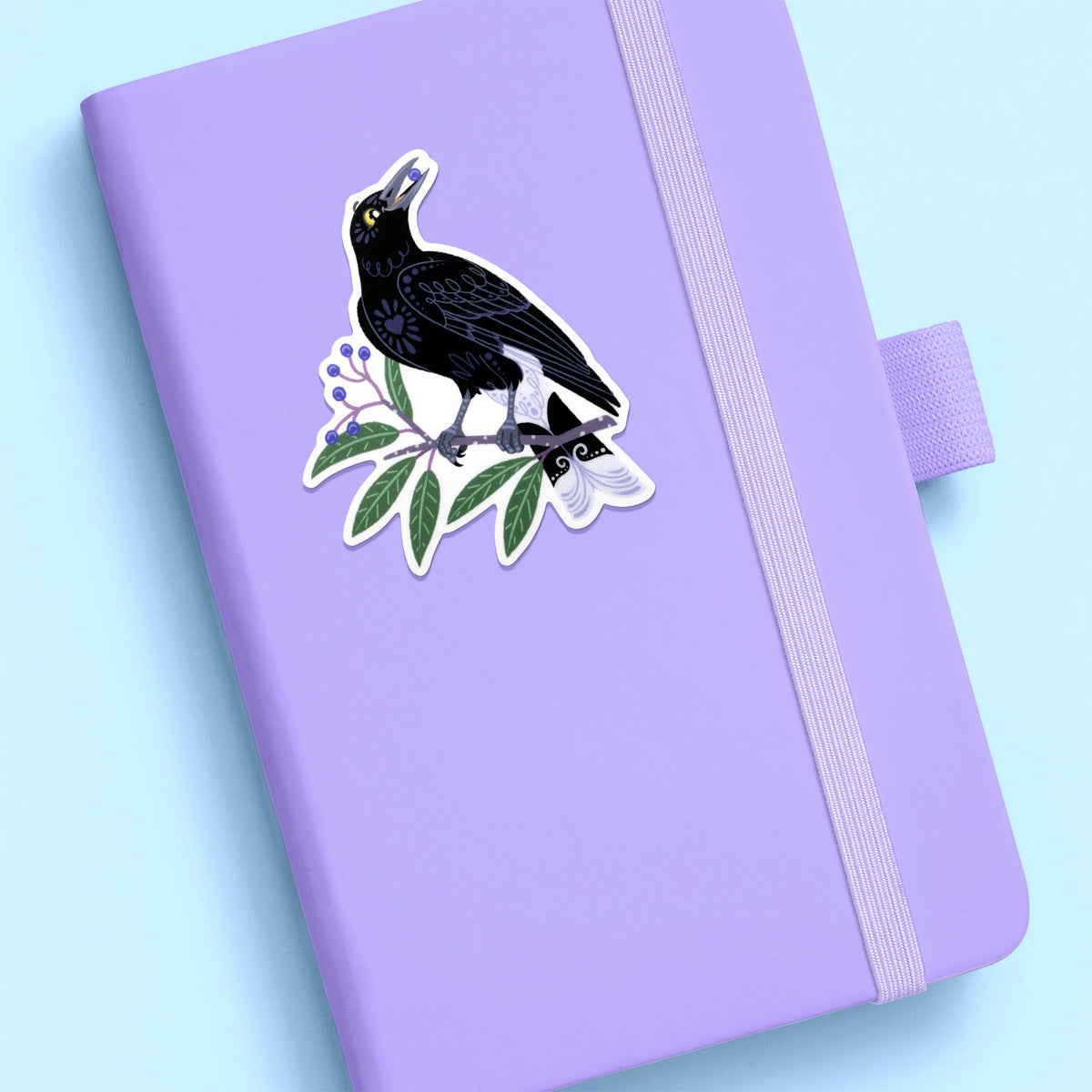Colourful diecut sticker of a pied currawong eating a blueberry ash berry stuck to a lilac journal on a pale blue background.