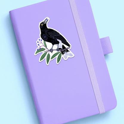 Colourful diecut sticker of a pied currawong eating a blueberry ash berry stuck to a lilac journal on a pale blue background.