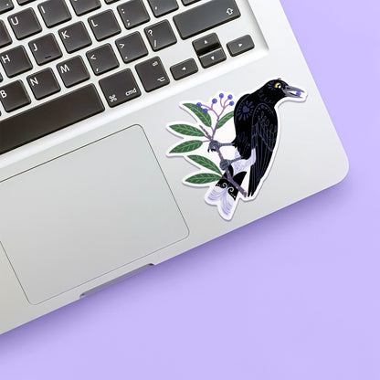 Colourful diecut sticker of a pied currawong eating a blueberry ash berry stuck below the keyboard of a Macbook laptop on a purple background.