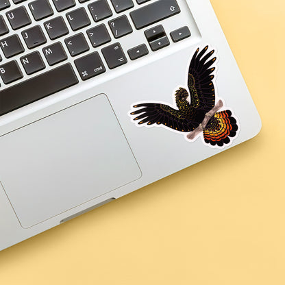 Colourful sticker of a female red-tailed black cockatoo perched on a branch with her wings out stuck below the keyboard of a Macbook laptop on yellow background.