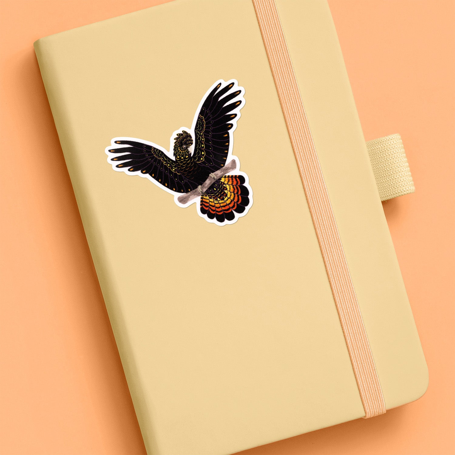 Colourful sticker of a female red-tailed black cockatoo perched on a branch with her wings out stuck to a pale yellow journal on an orange background.