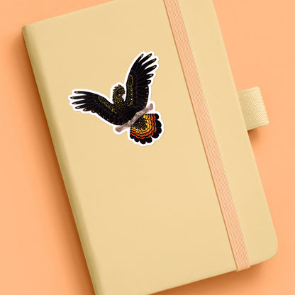 Colourful sticker of a female red-tailed black cockatoo perched on a branch with her wings out stuck to a pale yellow journal on an orange background.