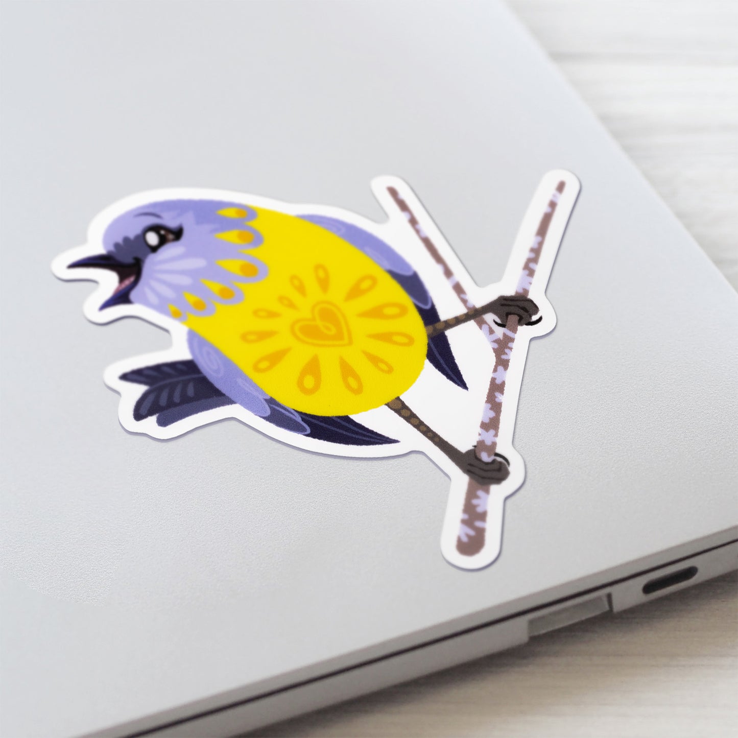 Closeup of a colourful sticker of an eastern yellow robin perched on a lichen-covered branch stuck to a lid of a Macbook laptop on a textured white wood background.