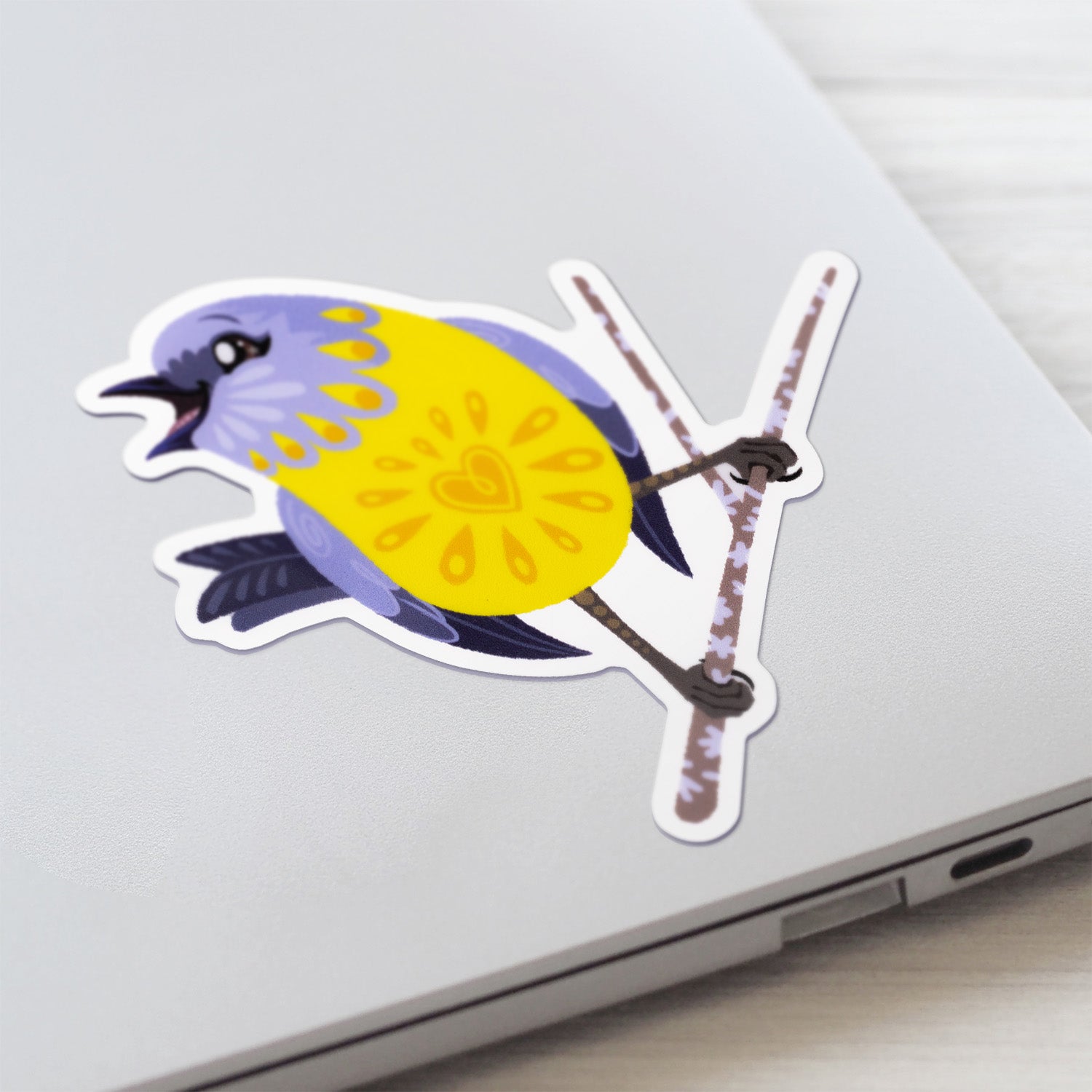 Closeup of a colourful sticker of an eastern yellow robin perched on a lichen-covered branch stuck to a lid of a Macbook laptop on a textured white wood background.