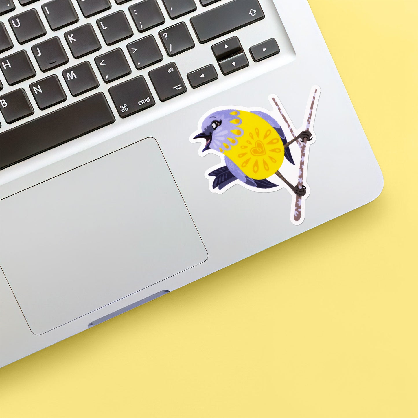 Colourful sticker of an eastern yellow robin perched on a lichen-covered branch stuck below the keyboard of a Macbook laptop on yellow background.
