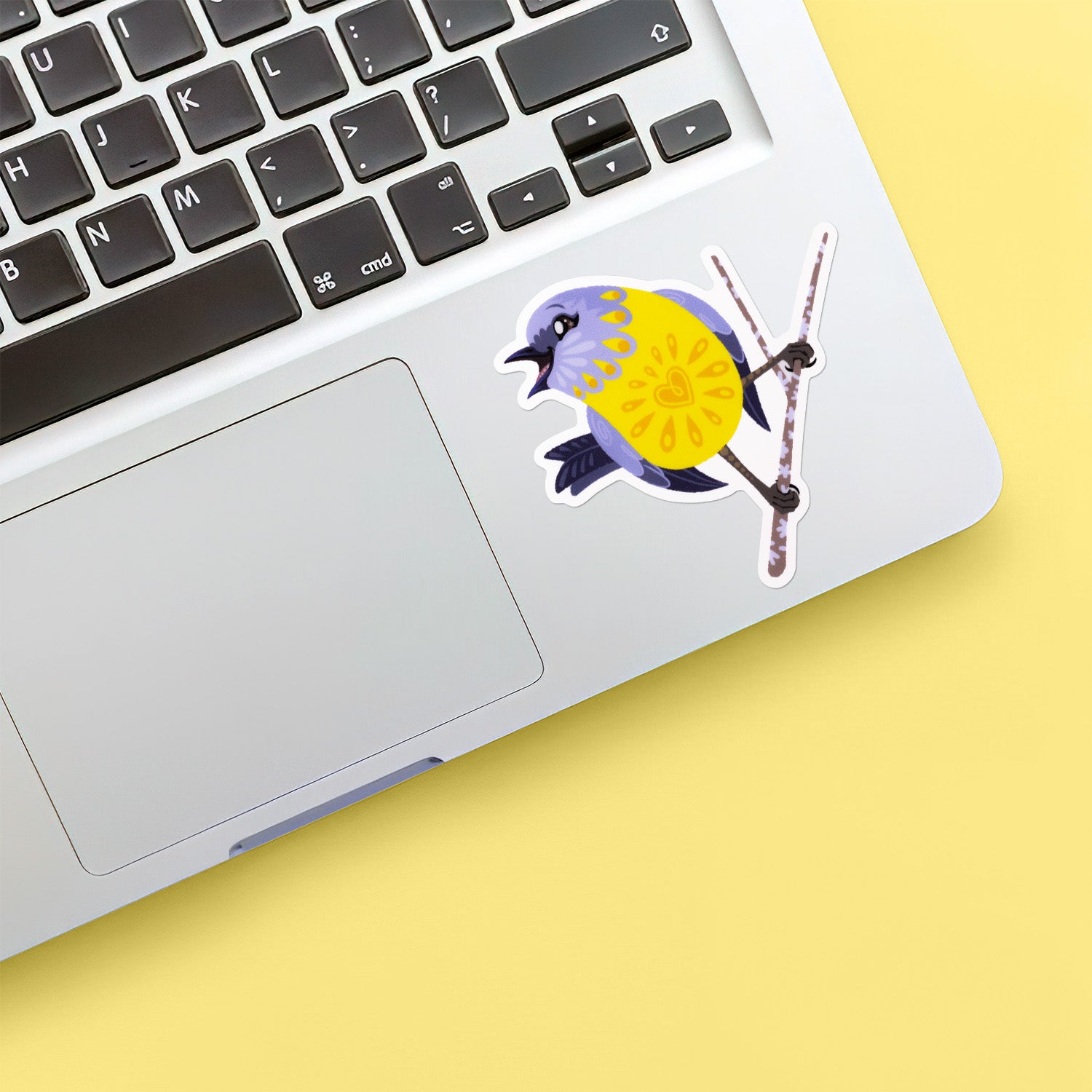 Colourful sticker of an eastern yellow robin perched on a lichen-covered branch stuck below the keyboard of a Macbook laptop on yellow background.