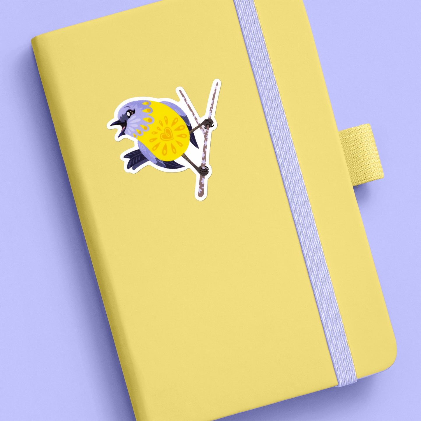 Colourful sticker of an eastern yellow robin perched on a lichen-covered branch stuck to a pale yellow journal on a lilac background.