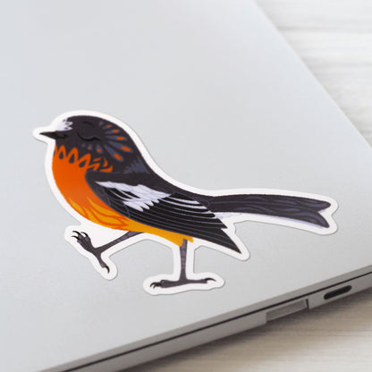 Closeup of a colourful sticker of a male flame robin walking with pep in his step stuck to a lid of a Macbook laptop on a textured white wood background.