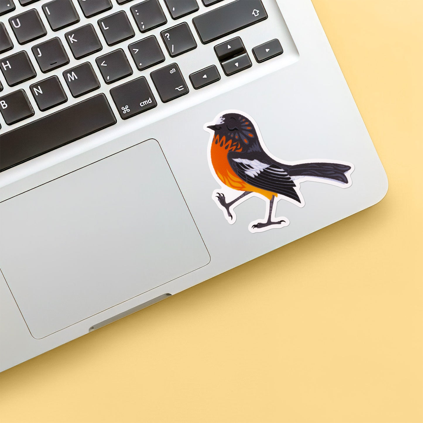 Colourful sticker of a male flame robin walking with pep in his step stuck below the keyboard of a Macbook laptop on a yellow background.