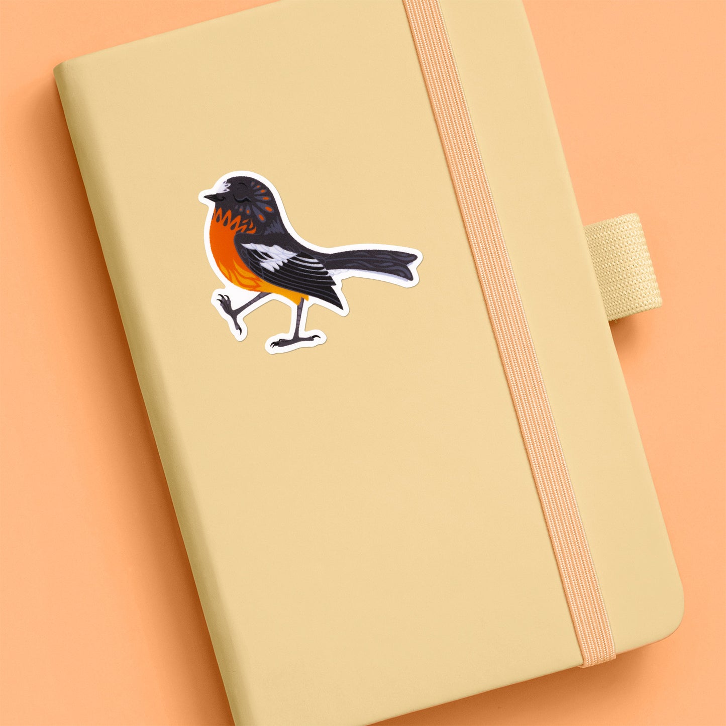Colourful sticker of a male flame robin walking with pep in his step stuck to a pale yellow journal on an orange background.