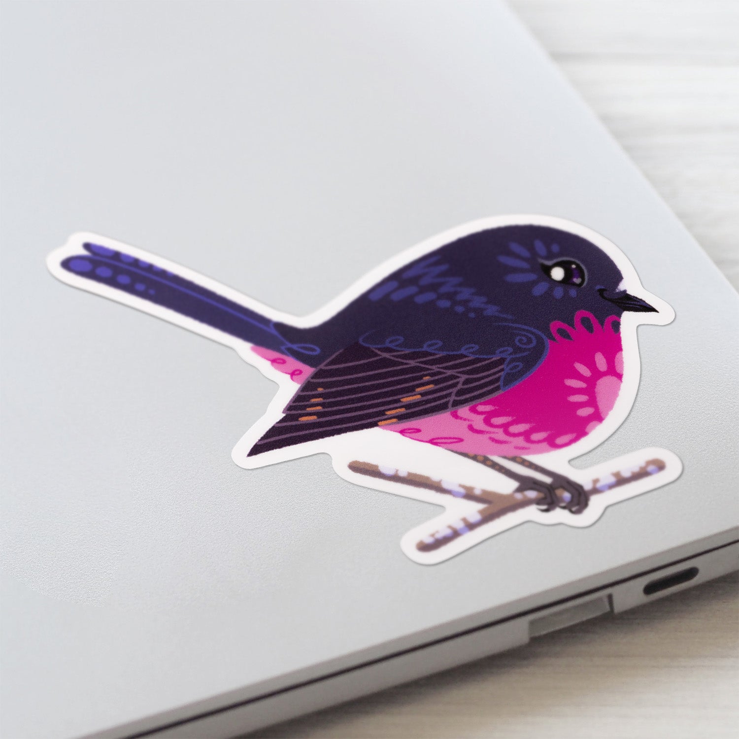 Closeup of a colourful sticker of a male pink robin perched on a lichen-covered branch stuck to a lid of a Macbook laptop on a textured white wood background.