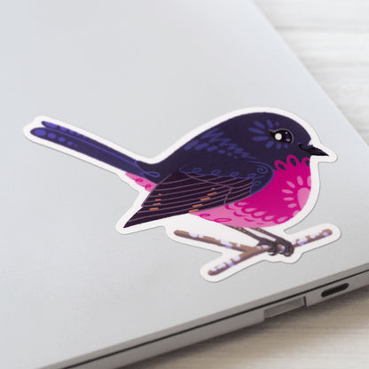 Closeup of a colourful sticker of a male pink robin perched on a lichen-covered branch stuck to a lid of a Macbook laptop on a textured white wood background.