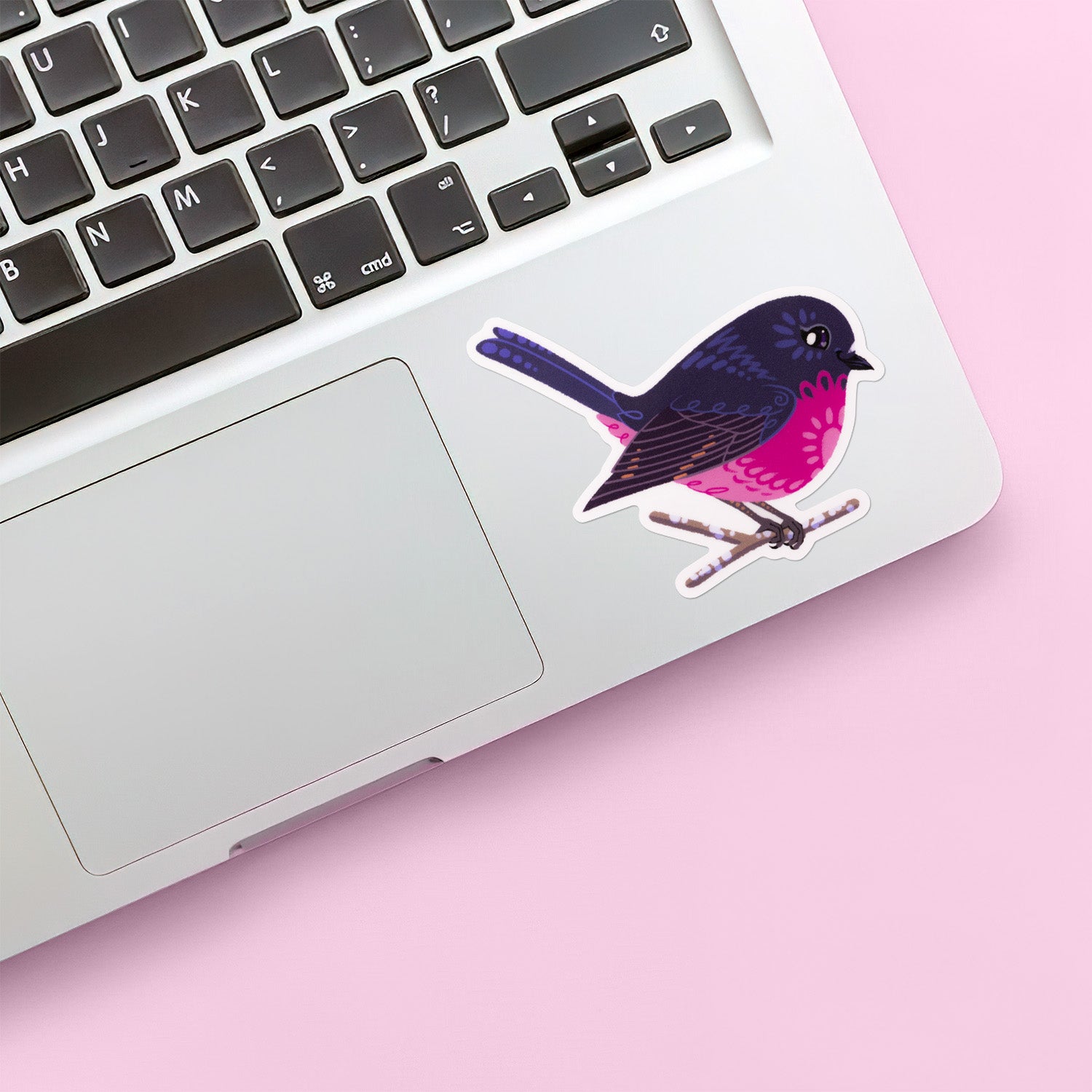 Colourful sticker of a male pink robin perched on a lichen-covered branch stuck below the keyboard of a Macbook laptop on light pink background.
