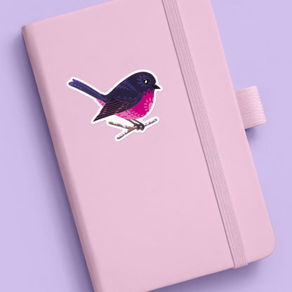 Colourful sticker of a male pink robin perched on a lichen-covered branch stuck to a pale pink journal on a lilac background.