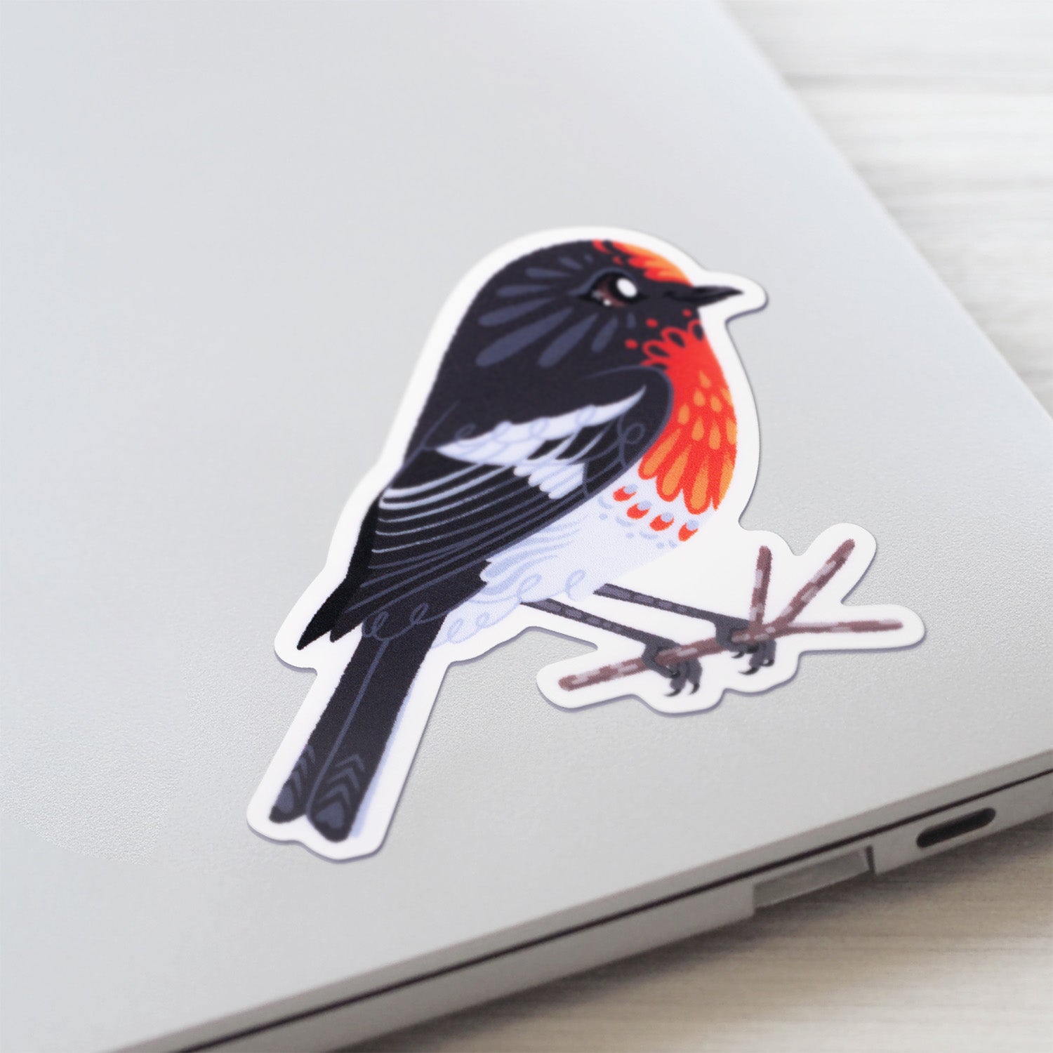Closeup of a colourful sticker of a male red-capped robin perched on a lichen-covered branch stuck to a lid of a Macbook laptop on a textured white wood background.