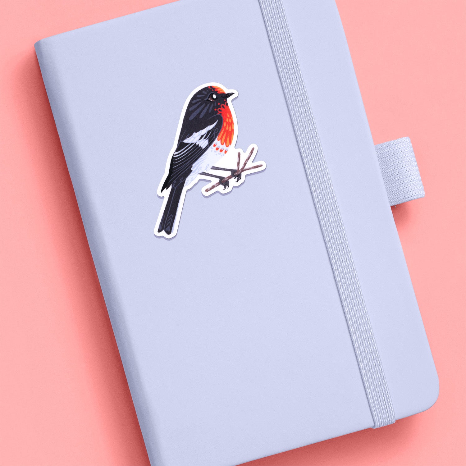 Colourful sticker of a male red-capped robin perched on a lichen-covered branch stuck to a pale grey journal on a rose pink background.