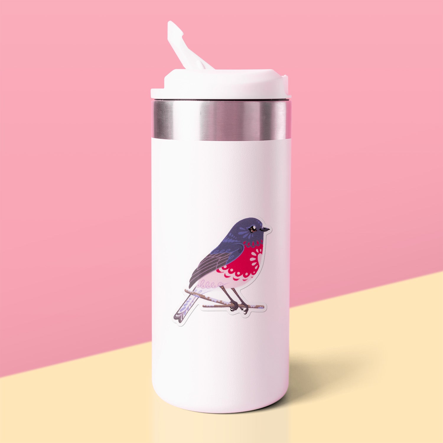 Colourful sticker of a male rose robin perched on a lichen-covered branch stuck to a white metal drink bottle on a yellow table on a pink background.