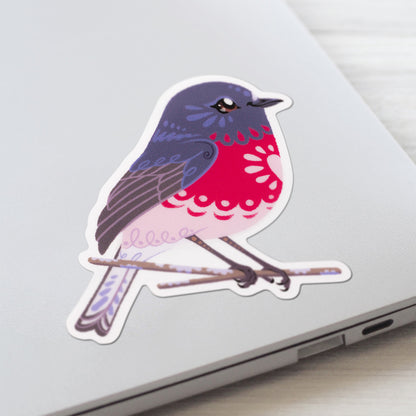 Closeup of a colourful sticker of a male rose robin perched on a lichen-covered branch stuck to a lid of a Macbook laptop on a textured white wood background.