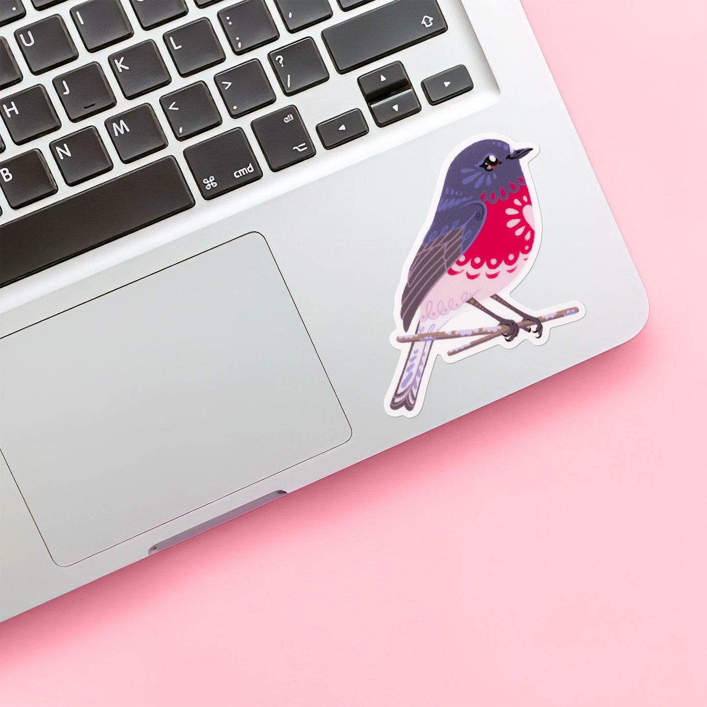 Colourful sticker of a male rose robin perched on a lichen-covered branch stuck below the keyboard of a Macbook laptop on pink background.