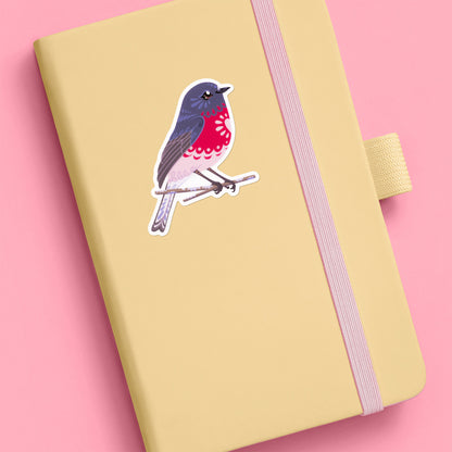 Colourful sticker of a male rose robin perched on a lichen-covered branch stuck to a pale yellow journal on a pink background.