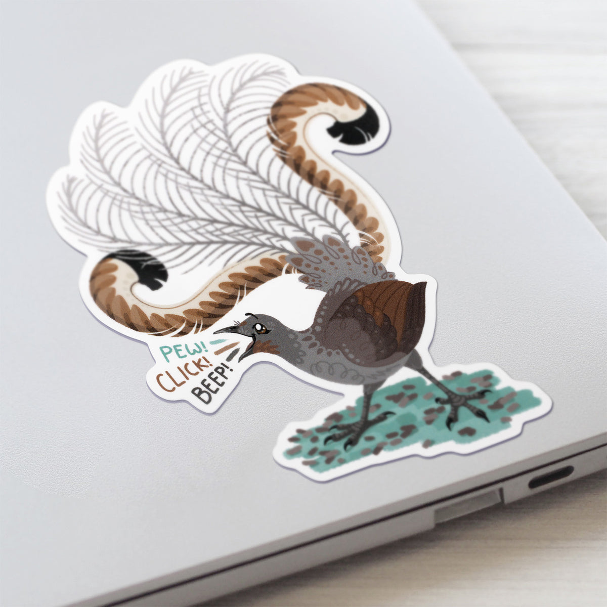 Closeup of a colourful sticker of a male superb lyrebird singing stuck to a lid of a Macbook laptop on a textured white wood background.