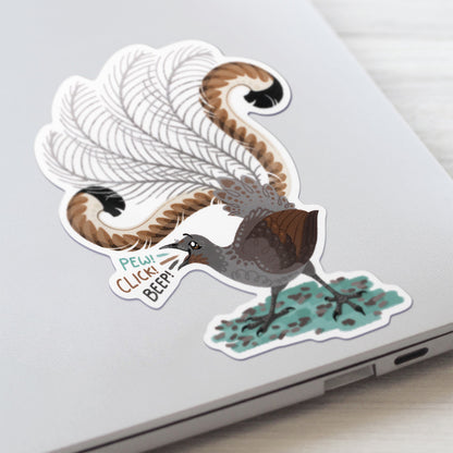 Closeup of a colourful sticker of a male superb lyrebird singing stuck to a lid of a Macbook laptop on a textured white wood background.