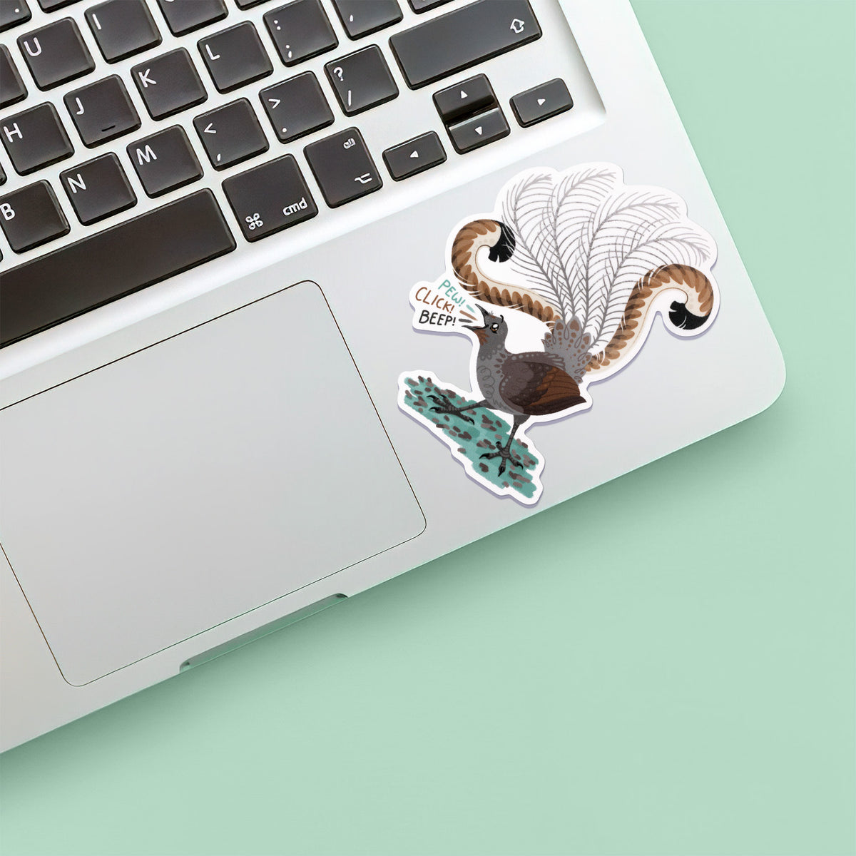 Colourful sticker of a male superb lyrebird singing stuck below the keyboard of a Macbook laptop on a pale green background.