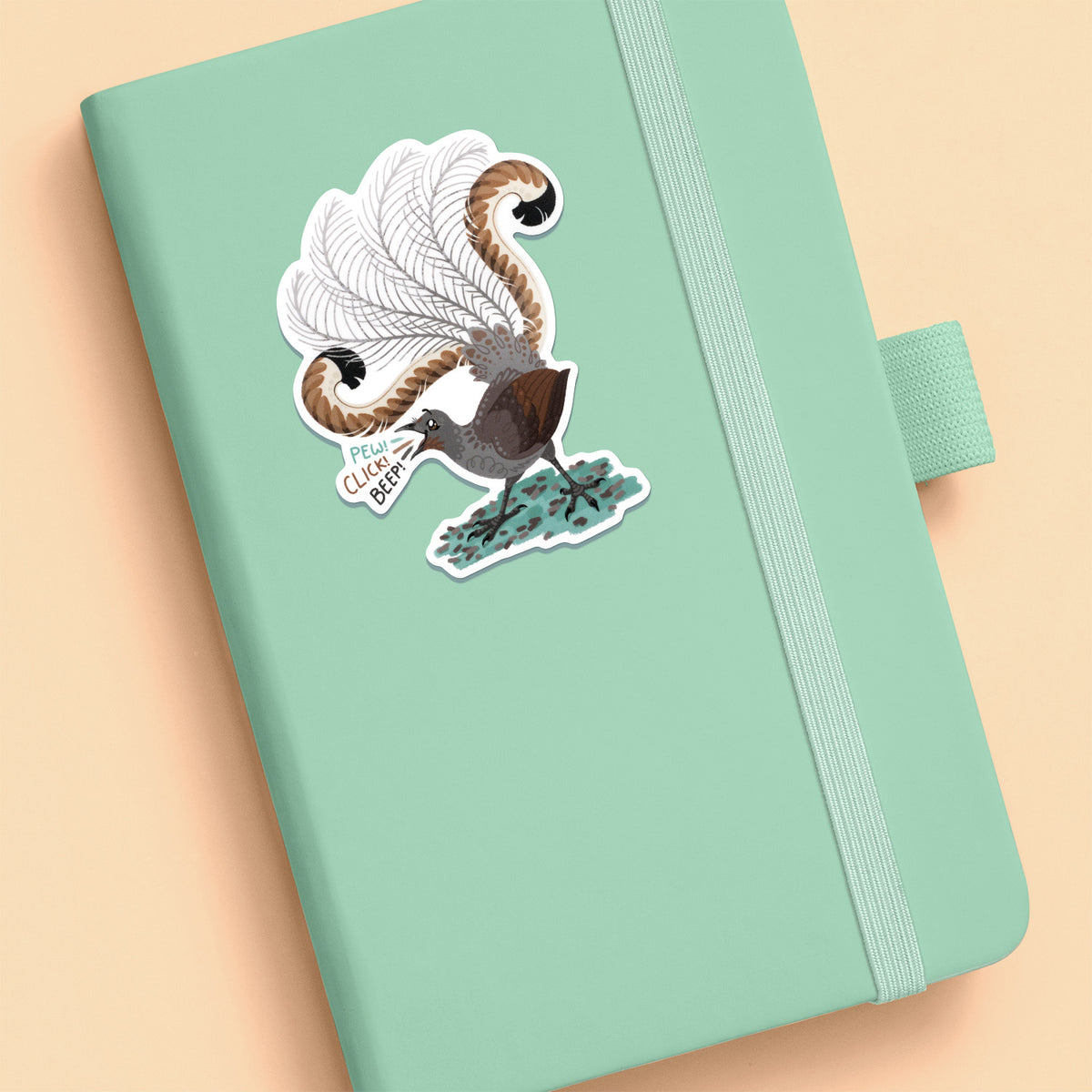 Colourful sticker of a male superb lyrebird singing stuck to a pale green journal on a beige background.