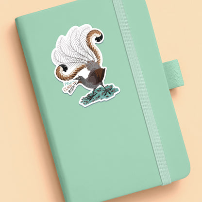 Colourful sticker of a male superb lyrebird singing stuck to a pale green journal on a beige background.
