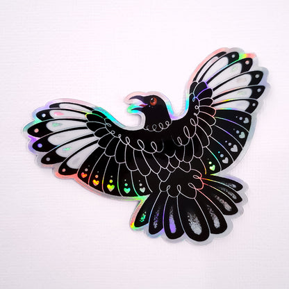 Real photo of a colourful holographic metallic die-cut sticker of a white-winged chough with spread wings on white canvas texture background.