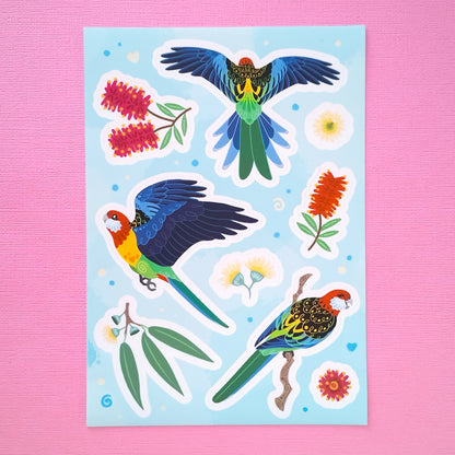 Real photo of a aolourful A5 sticker sheet of 3 different Eastern Rosellas, eucalyptus leaves, eucalyptus flowers and red & pink bottlebrush flowers on a pink canvas texture background.