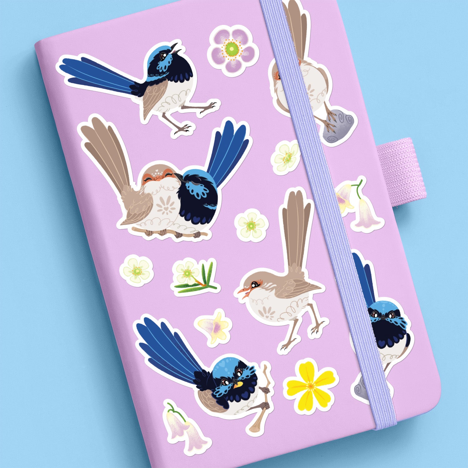 Colourful stickers of male & female Superb Fairywrens and assorted native flowers stuck to a lilac journal on a blue background.