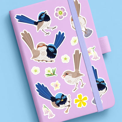 Colourful stickers of male & female Superb Fairywrens and assorted native flowers stuck to a lilac journal on a blue background.