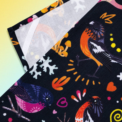 Closeup of a hanging strap on the reverse side of a tea towel with a black, pink, orange and yellow Australian robins pattern sitting flat on a pastel yellow and lilac background.