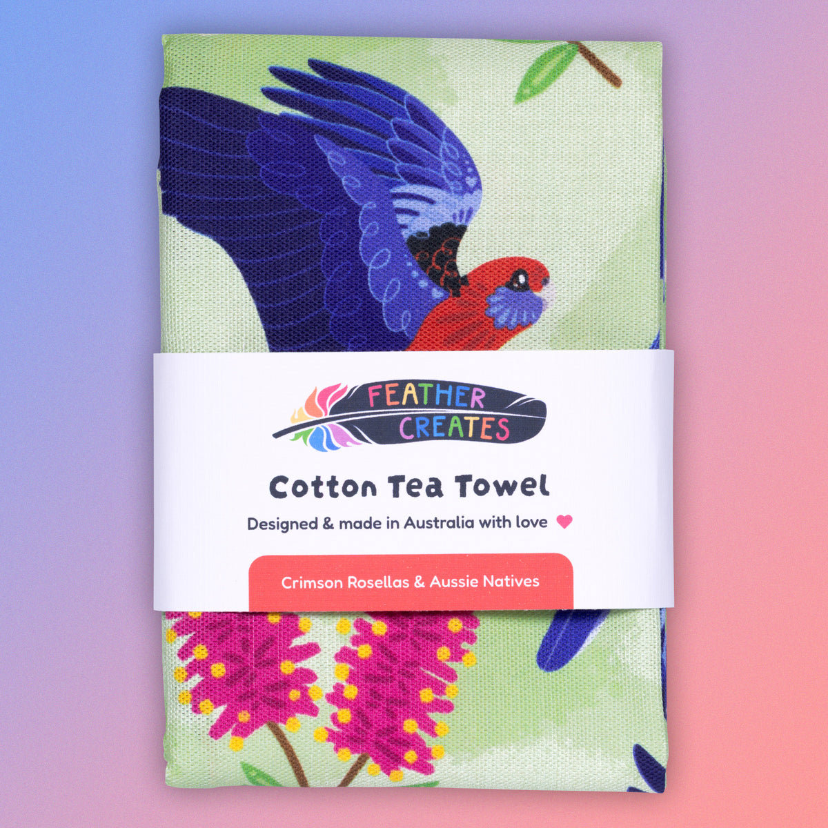 A folded tea towel with a green, red and blue crimson rosellas pattern sitting flat on a pastel blue and pink background.