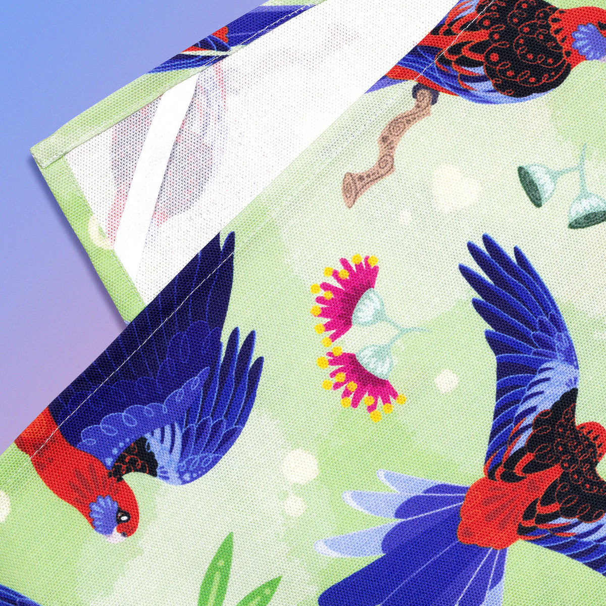 Closeup of a hanging strap on the reverse side of a tea towel with a green, red and blue crimson rosellas pattern sitting flat on a pastel blue and pink background.