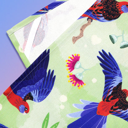 Closeup of a hanging strap on the reverse side of a tea towel with a green, red and blue crimson rosellas pattern sitting flat on a pastel blue and pink background.