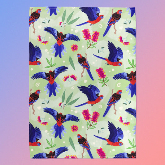 A tea towel with a green, red and blue crimson rosellas pattern sitting flat on a pastel blue and pink background.