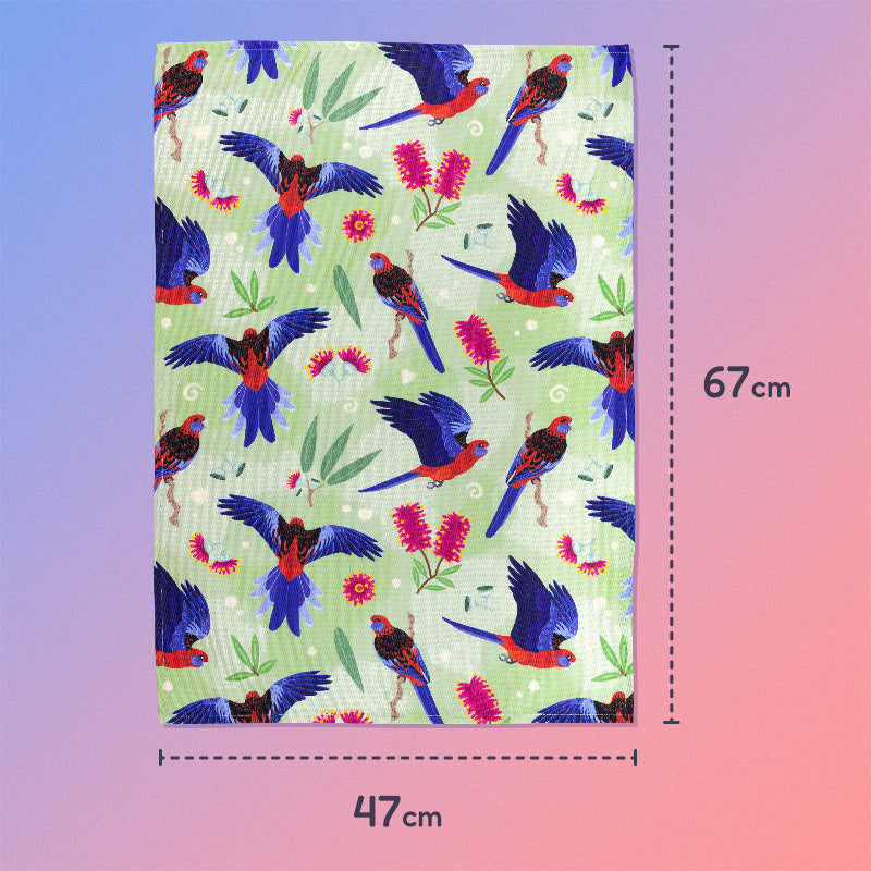 47x67cm tea towel with a green, red and blue crimson rosellas pattern sitting flat on a pastel blue and pink background.