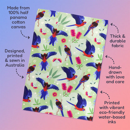 Features & benefits ('Made from 100% half panama cotton canvas, designed, printed & sewn in Australia, thick & durable fabric, hand-drawn with love and care, printed with vibrant eco-friendly water-based inks) surrounding a tea towel with a green, red and blue crimson rosellas pattern sitting flat on a pastel blue and pink background.