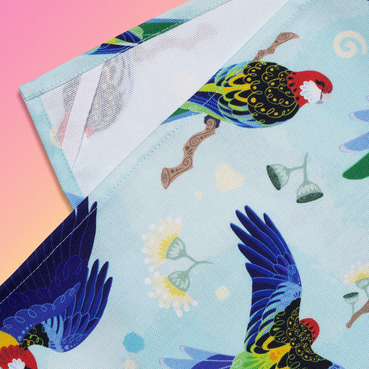 Closeup of a hanging strap on the reverse side of a tea towel with a rainbow eastern rosellas pattern sitting flat on a pastel pink, yellow and green background.