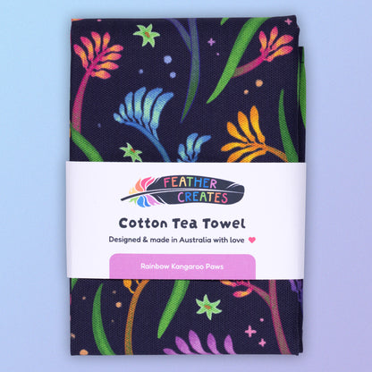 A folded tea towel with a rainbow kangaroo paw pattern sitting on a pastel blue and purple background.