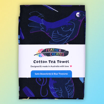 A folded tea towel with a black, blue, purple and yellow satin bowerbirds pattern sitting flat on a pastel yellow and blue background.