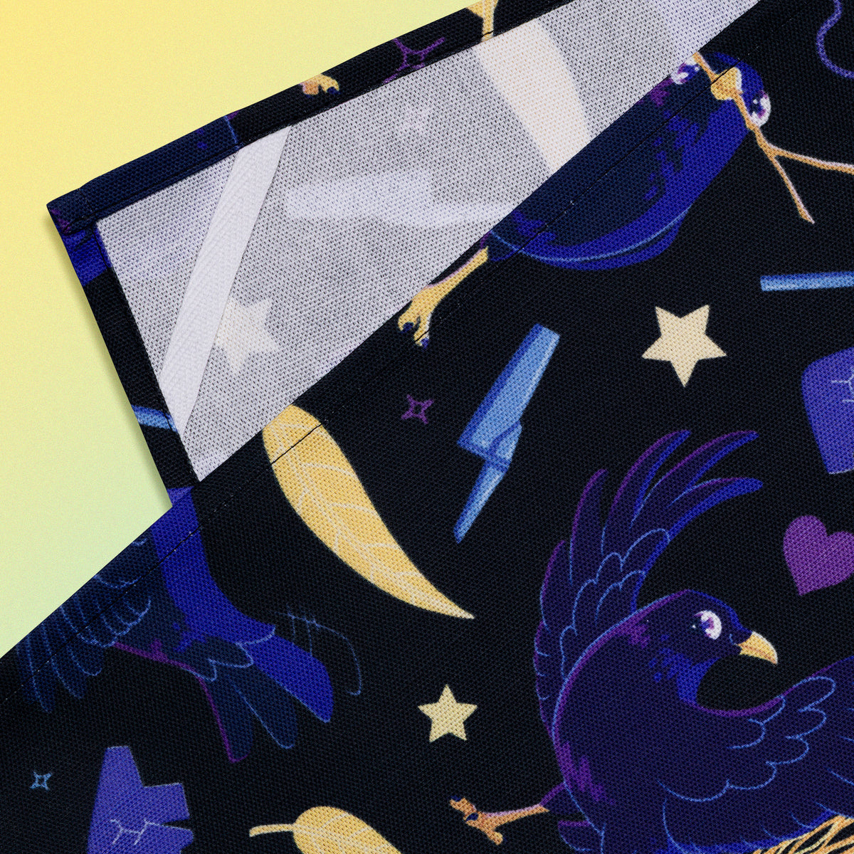 Closeup of a hanging strap on the reverse side of a tea towel with a black, blue, purple and yellow satin bowerbirds pattern sitting flat on a pastel yellow and blue background.
