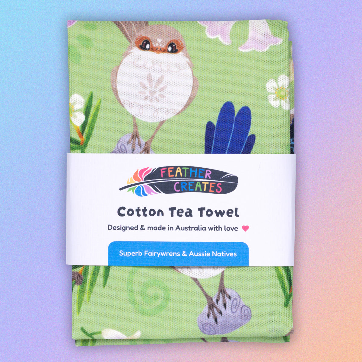 A folded tea towel with a green, blue, yellow and beige superb fairywrens pattern sitting flat on a pastel blue, pink and apricot background.
